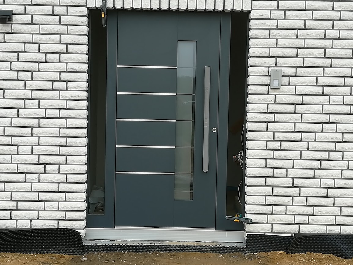 Moderne graue Haustür mit vertikalem Glasstreifen, horizontalen Metallleisten vor weißer Ziegelwand.