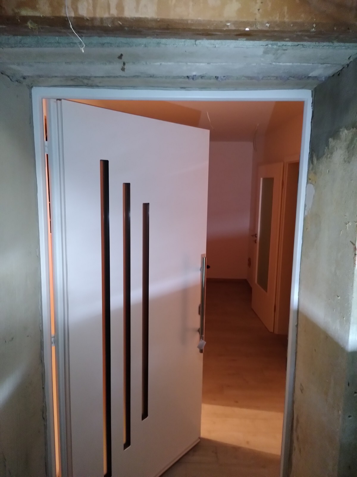 Offene weiße Tür mit drei vertikalen schwarzen Glasfenstern; Blick in Flur mit Holzfußboden.