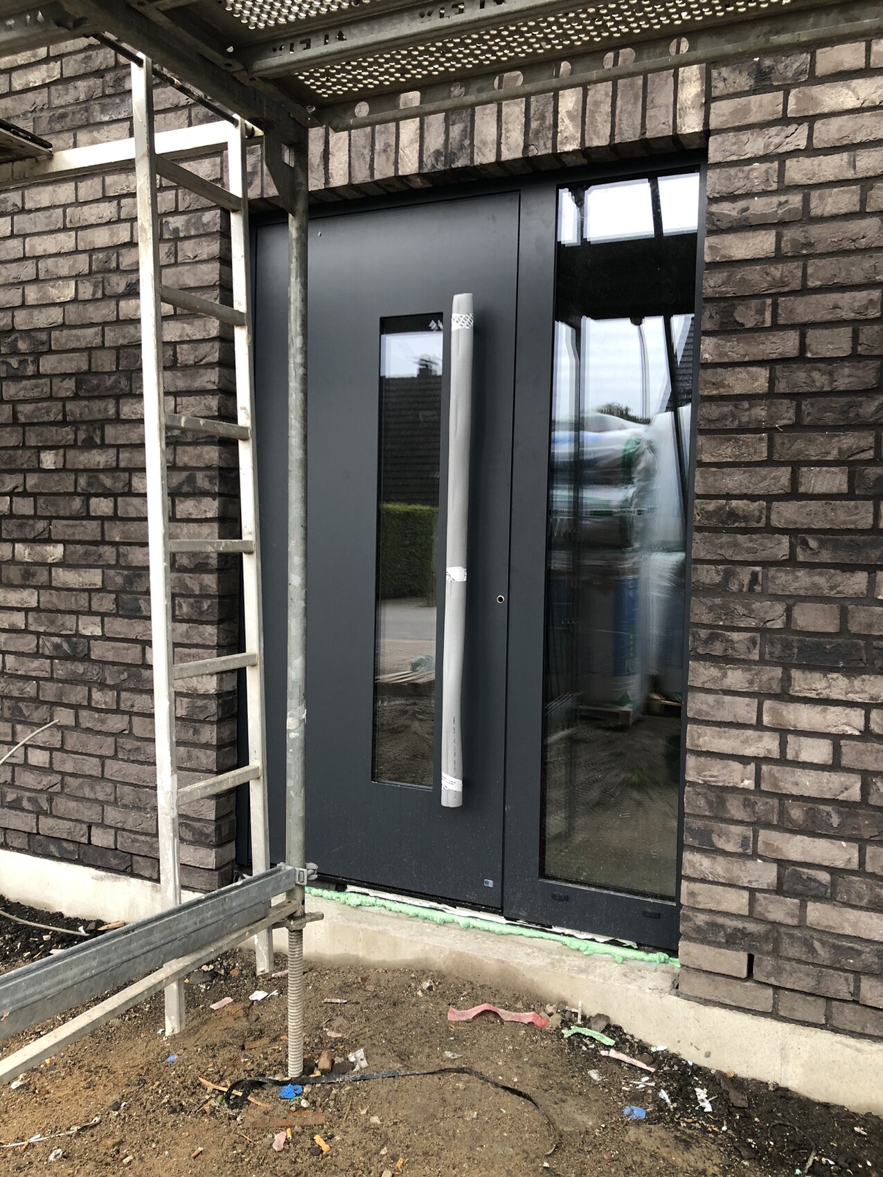 Moderne graue Haustür mit vertikalem Glasfenster vor Backsteinwand; Baugerüst davor.
