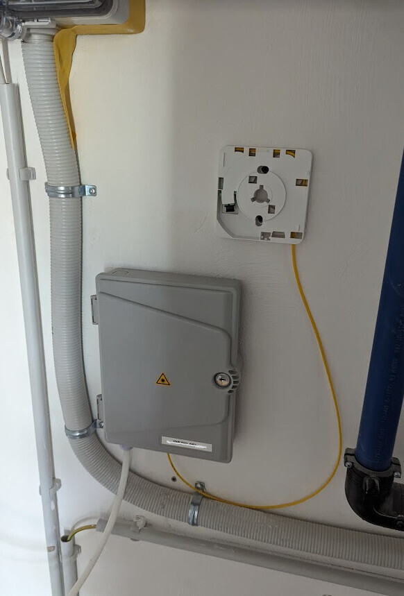 Wandmontiertes graues Elektro-Gehäuse mit Warnsymbol, gelber Zuleitung und Kabelkanäle.