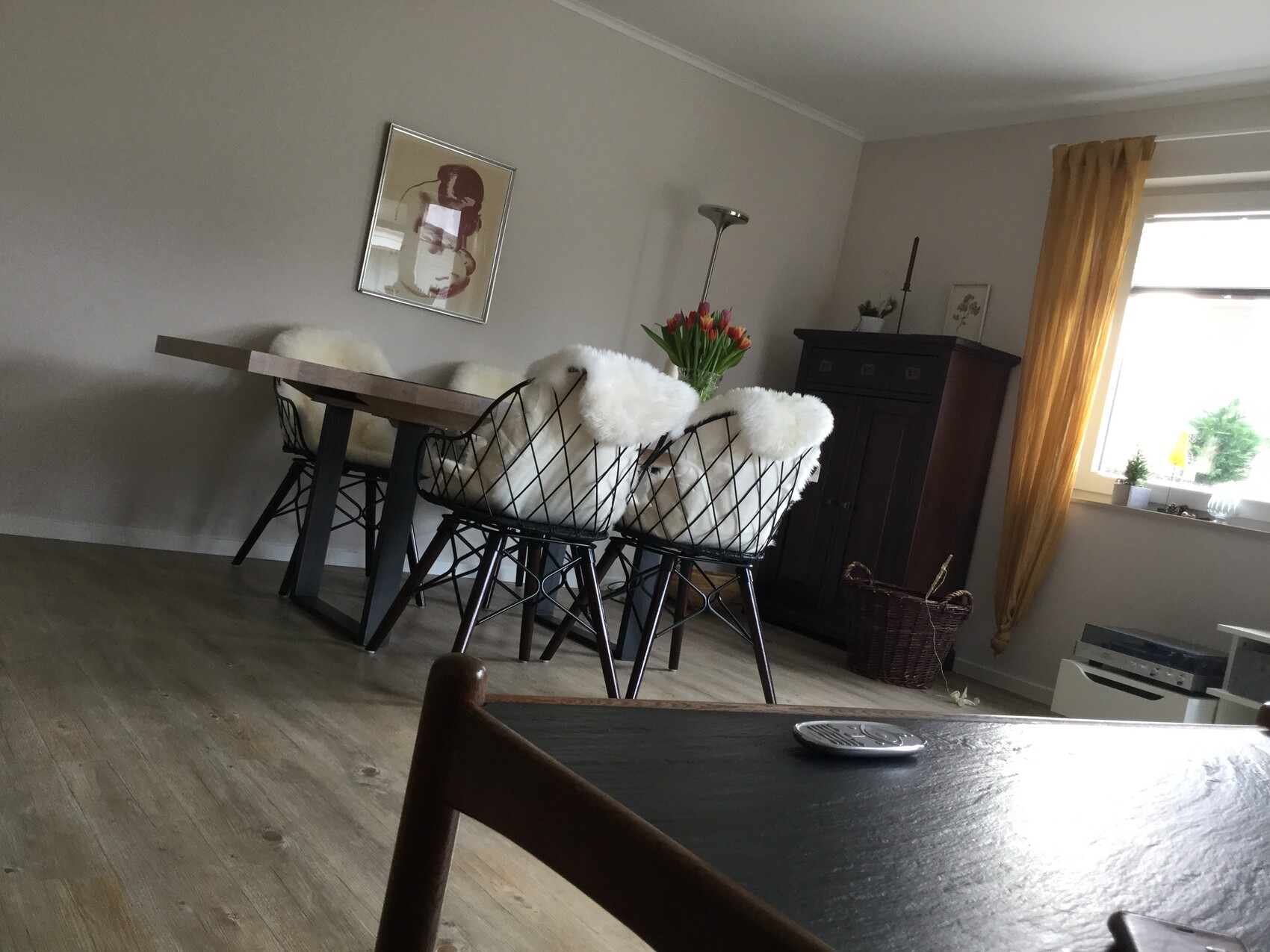 Wohnzimmer mit Esstisch, Stühlen mit weißen Fellauflagen, Tulpen, dunkler Schrank, goldene Vorhänge.