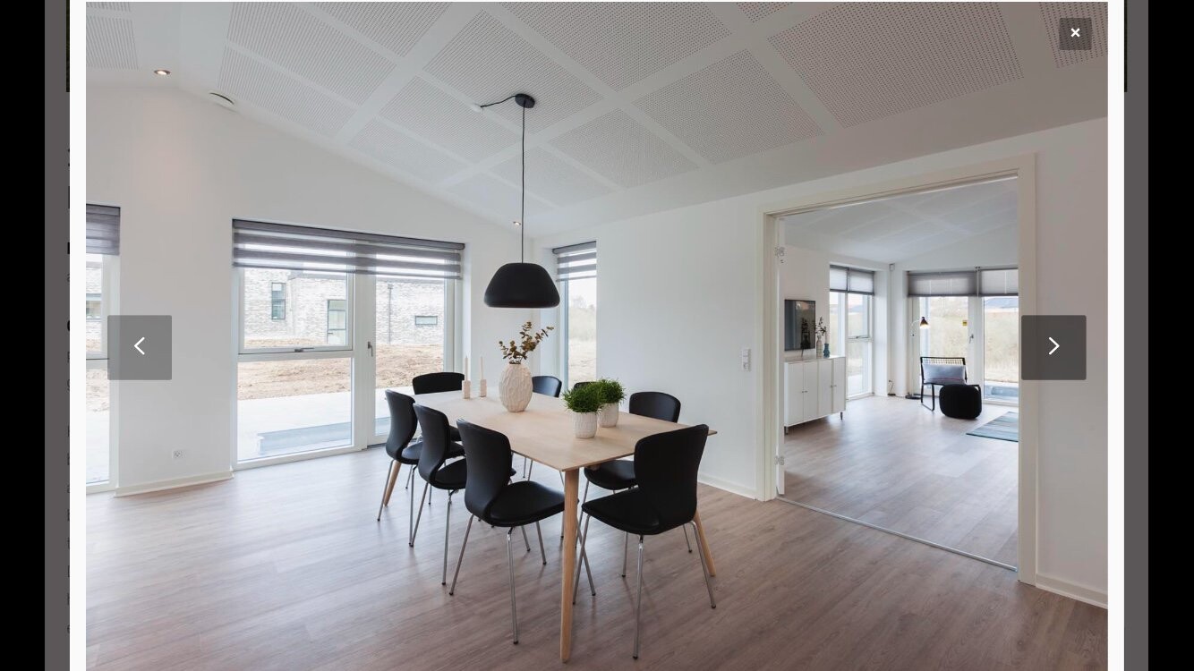 Helles Esszimmer mit Holztisch, sechs schwarzen Stühlen, großen Fenstern und offener Wohnbereich.