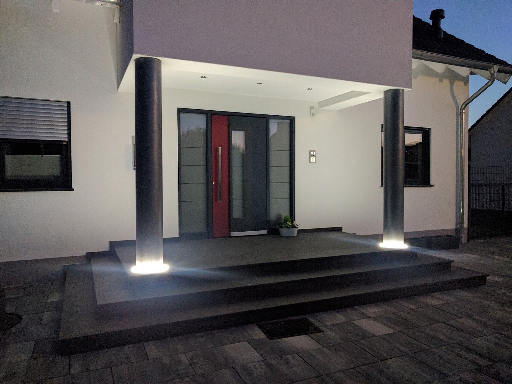 Moderne Frontansicht eines Hauses mit Eingangsbereich, zwei Saeulen und beleuchteten Stufen.