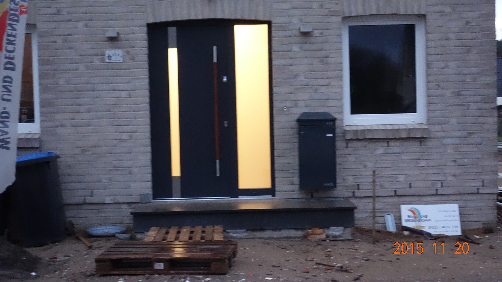 Moderne schwarze Eingangstür mit vertikalen Lichtstreifen vor Ziegelwand. Baustellenszene vor Hausfront.