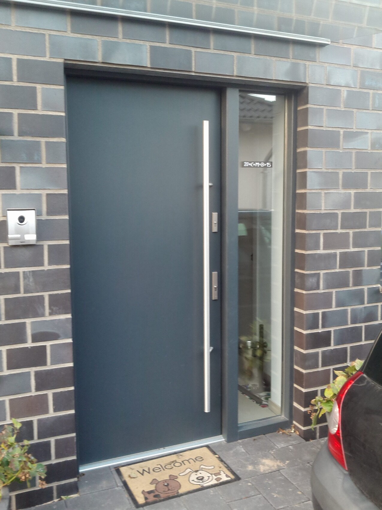 Moderne dunkle Haustür mit langem Metallgriff neben Glasfront; Willkommensmatte mit Tiermotiven.