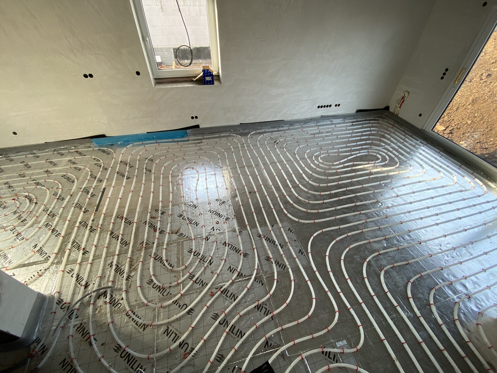 Verlegte Fußbodenheizrohre in spiralförmigem Muster auf Dämmfolie in einem Bauzimmer mit Fenster.