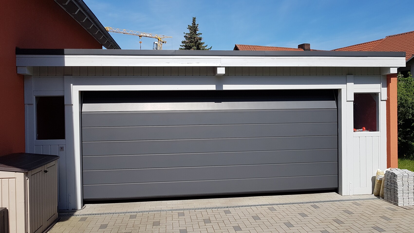 Garage mit grauem Sektionaltor, weißem Rahmen; orange Wand links, Pflastersteine rechts.
