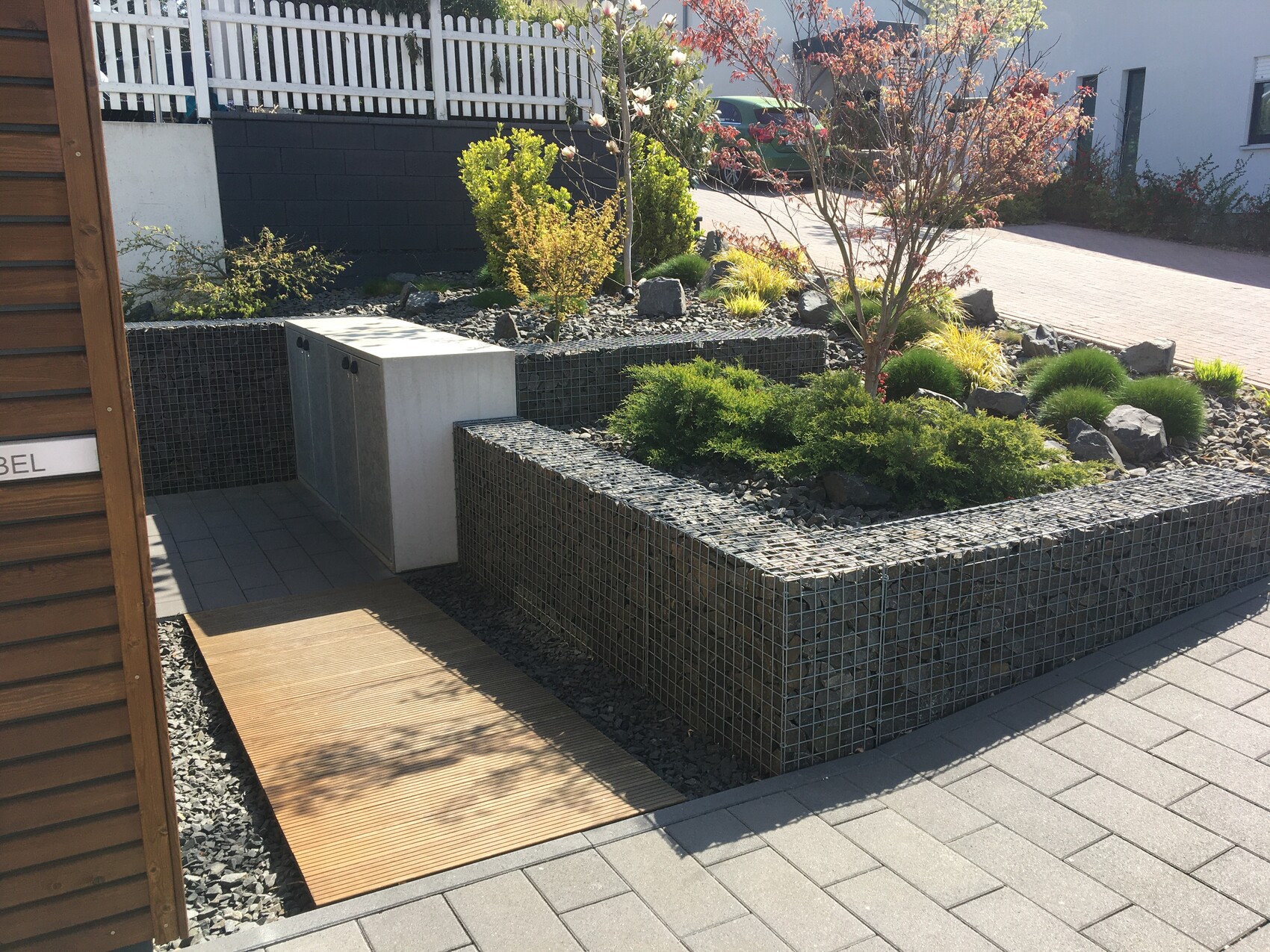 Moderne Gartenanlage mit Gabionenmauern, Steinbeeten, Bäumen und gepflastertem Weg.