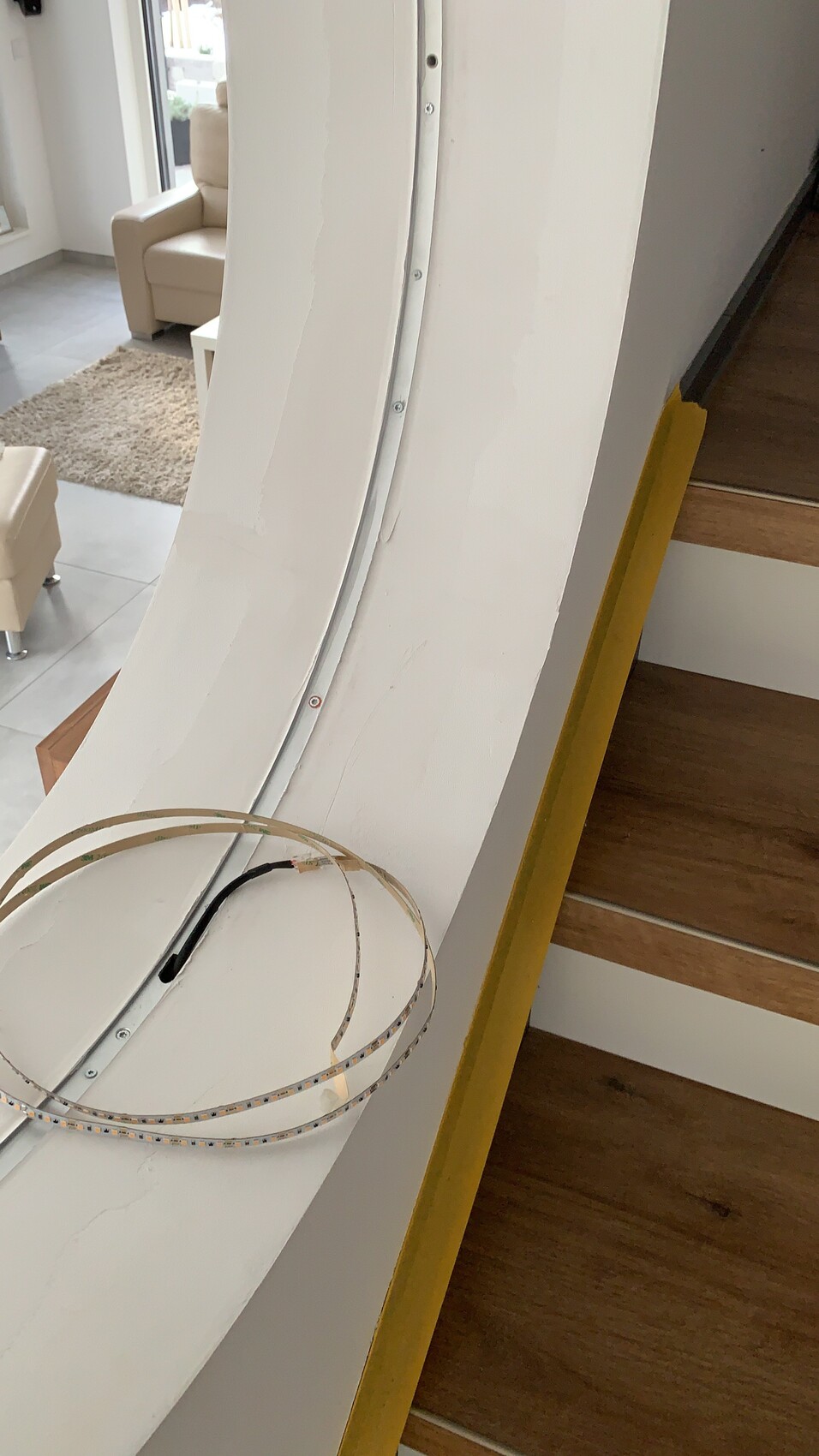 Weiße, gekrümmte Wand mit LED-Lichtband; Treppe mit gelbem Rand, Wohnzimmer dahinter.