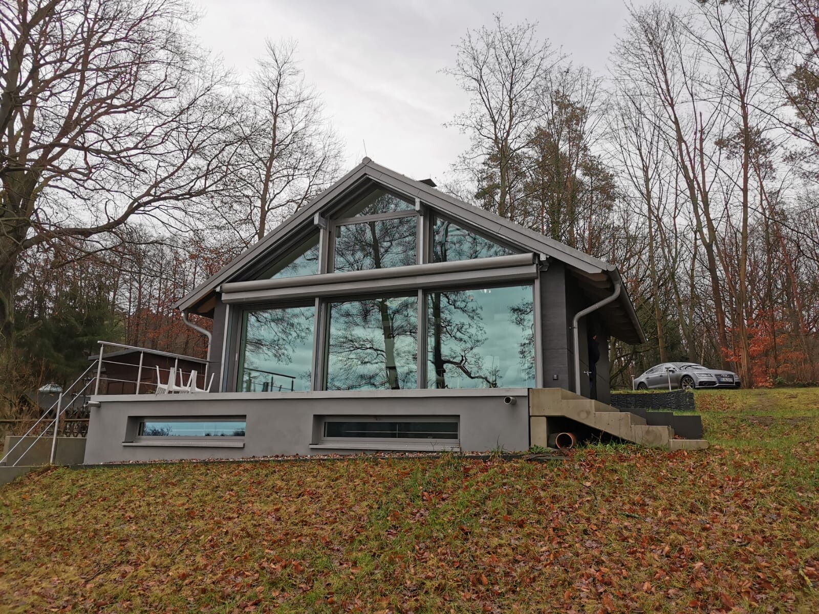 Moderne Glasfront eines Hauses am Hang, umgeben von Bäumen und Herbstlaub; Auto rechts.