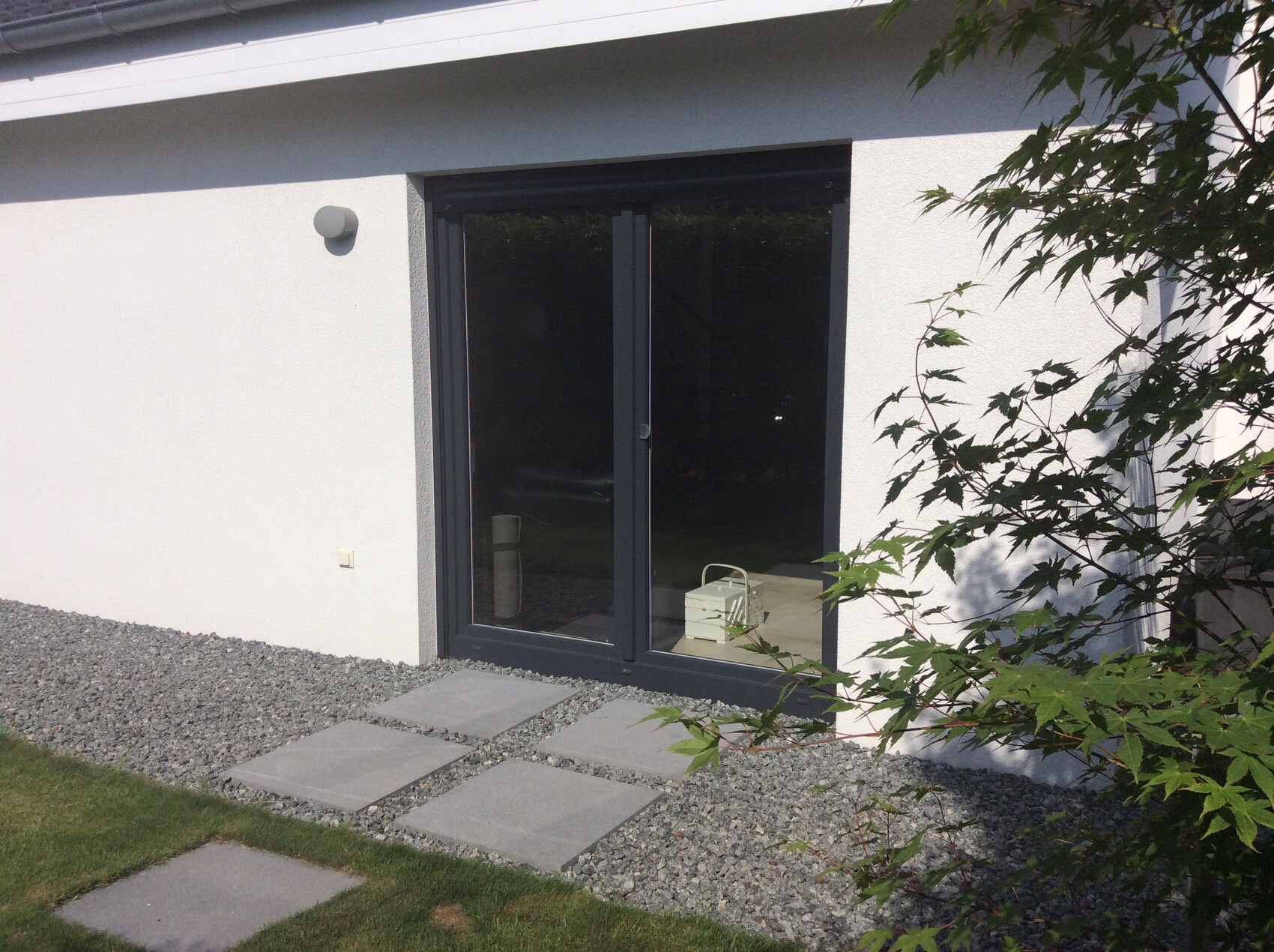 Weiße Hausfassade mit dunkler Glasschiebetür; Steinplattenweg auf Kies, grüne Pflanzen rechts.