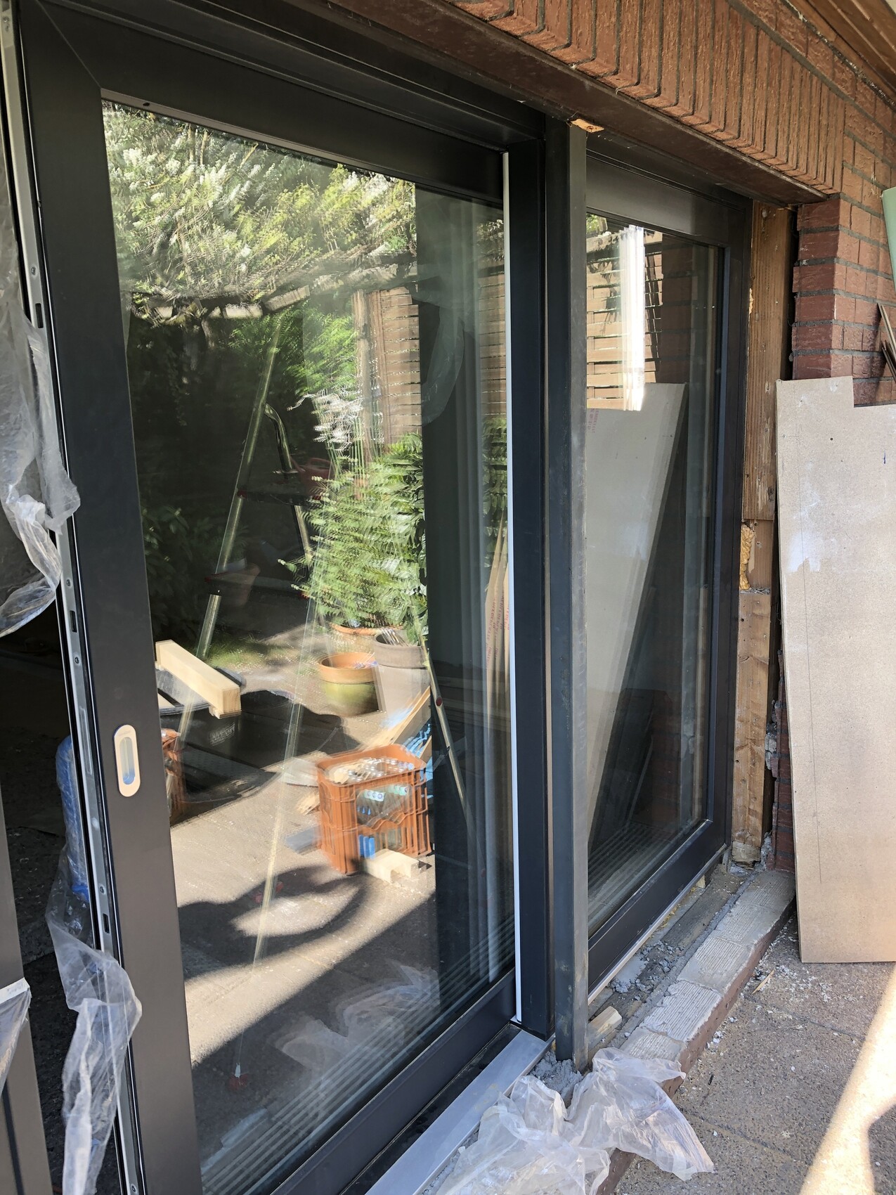 Großes Glas-Schiebetor vor Bauwand; Leiter, Bauholz, Pflanzen und Abdeckmaterial.