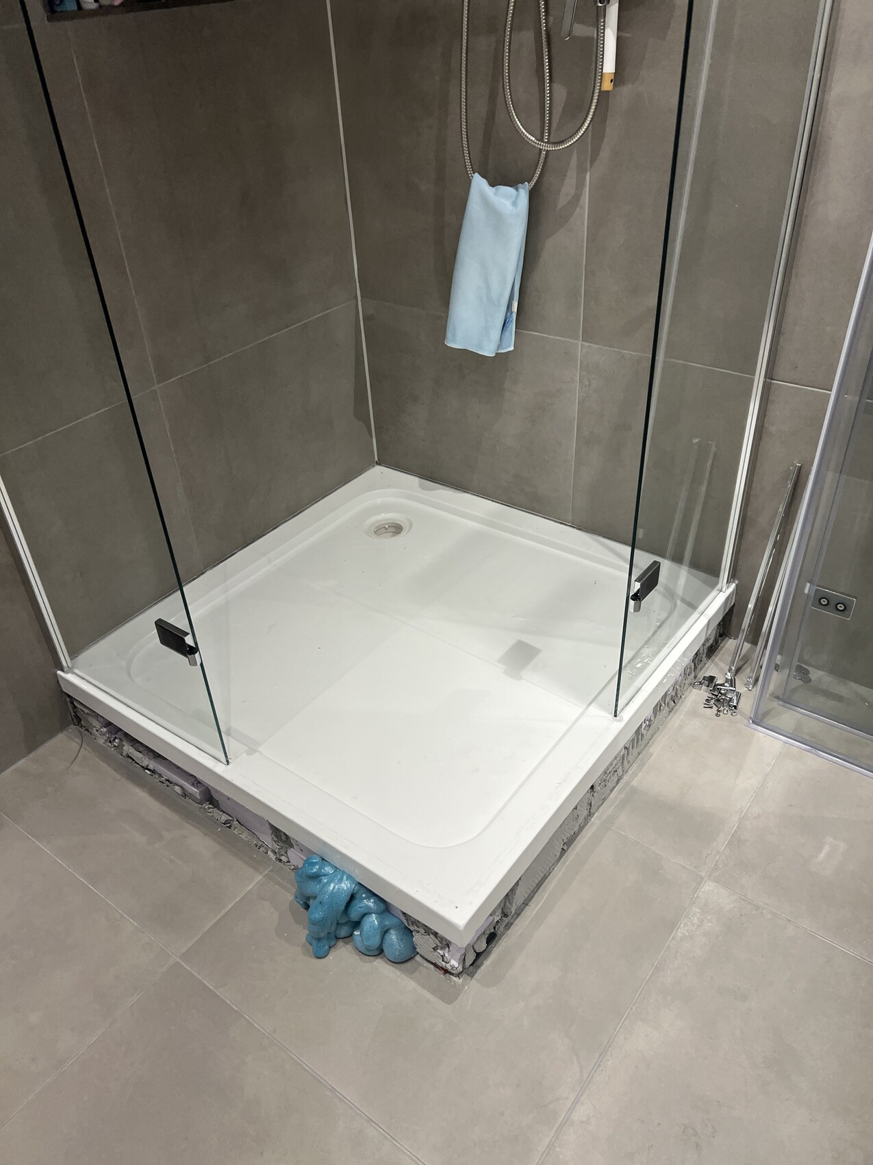 Badezimmer-Dusche mit Glaswänden, grauen Fliesen, weiße Duschwanne, Handbrause und blauem Handtuch.