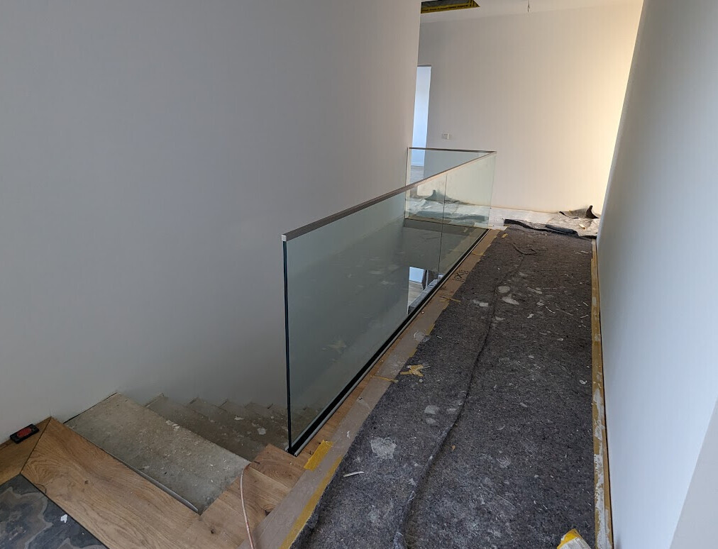 Innenraum eines Bauprojekts: Glasgeländer entlang Treppenöffnung, unfertiger Boden.