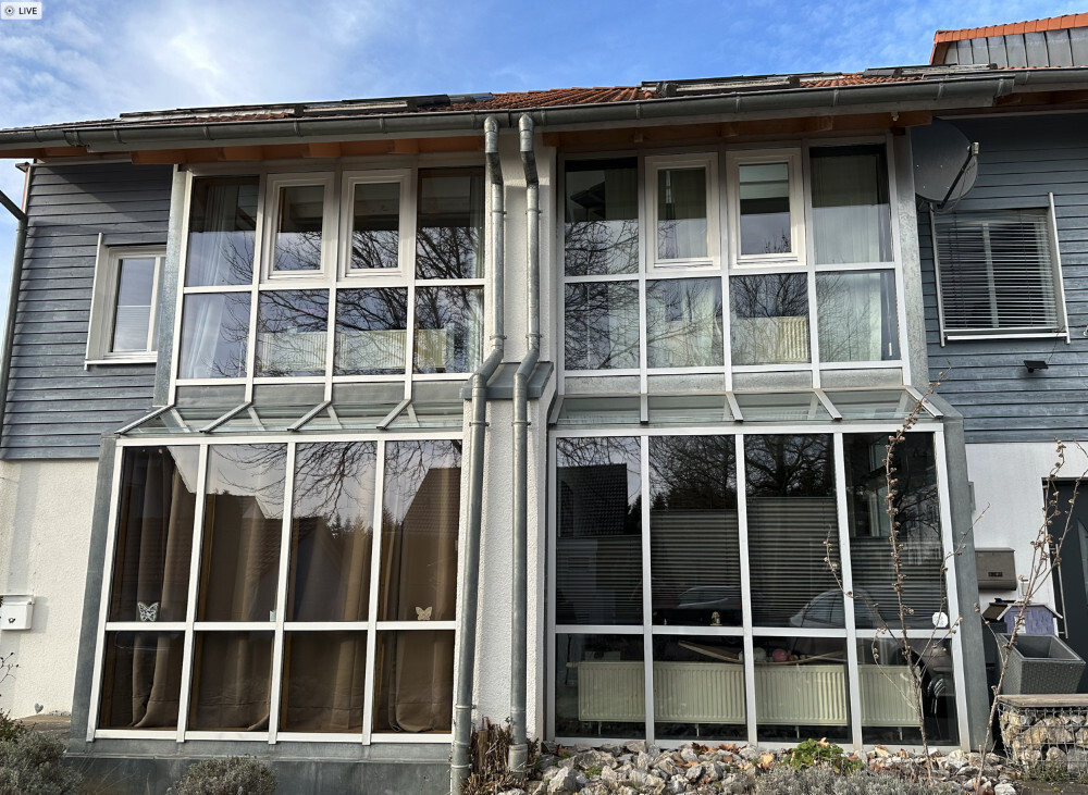Zweistöckiges Haus mit großen Glasfronten und grauen Metallrahmen, Fenstervorhänge sichtbar.
