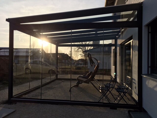 Glas-Wintergarten am Haus mit schwarzem Metallrahmen, Gartenstühle, Sonnenuntergang im Hintergrund