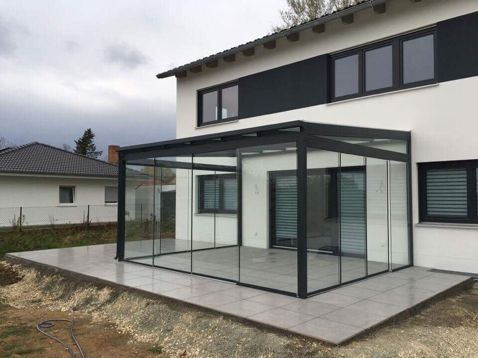 Moderne Glasveranda mit schwarzem Metallrahmen an weißem Haus, große Glasfront und Terrasse.