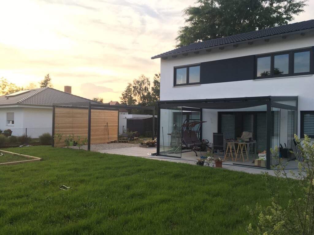 Moderne weiße Hausfront mit glasüberdachter Veranda und Gartenmöbeln, Abendlicht