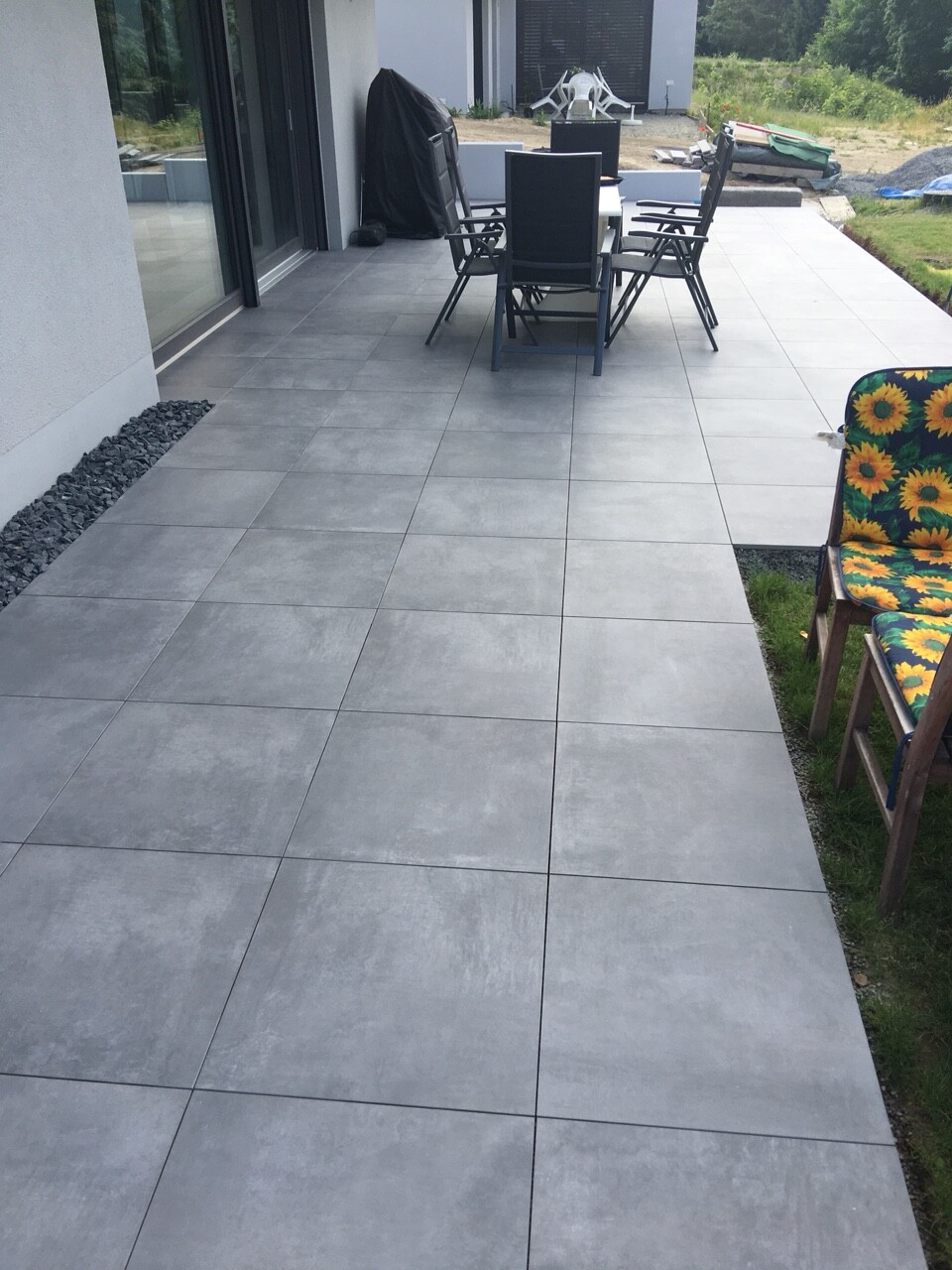 Große graue Fliesen-Terrasse mit Esstisch und Stühle vor dem Haus; Sonnenblumen-Stuhl am Rand.