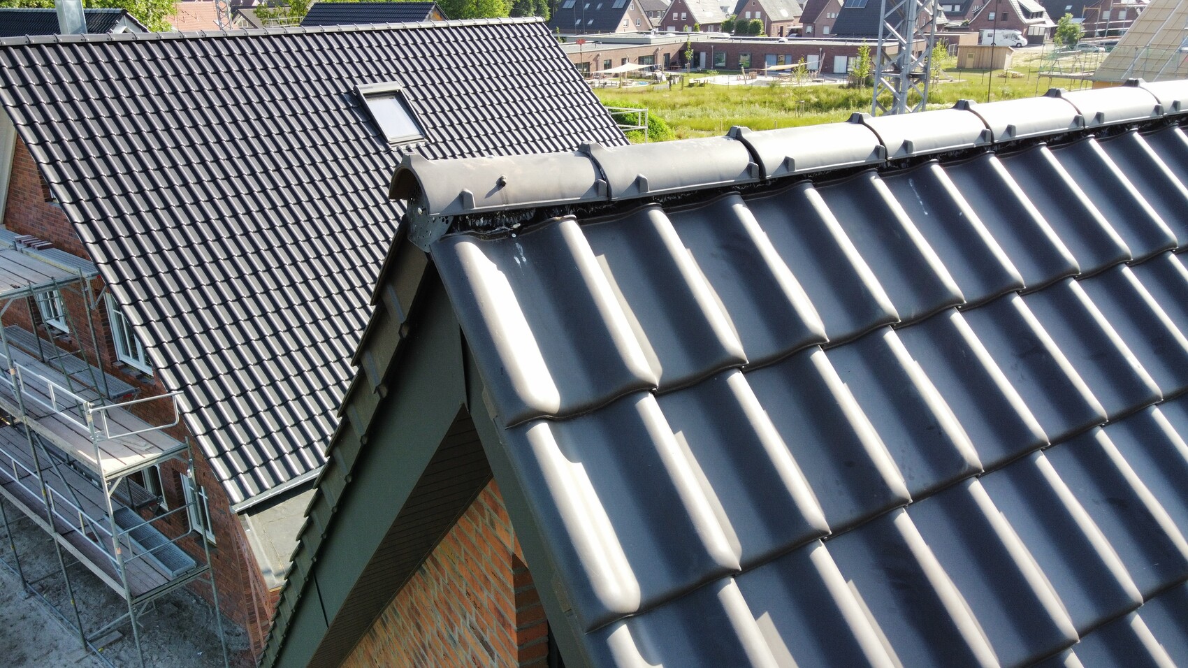 Blick von oben auf dunkle Dachziegel eines Neubauhauses mit Gerüst und weiteren Häusern im Hintergrund.