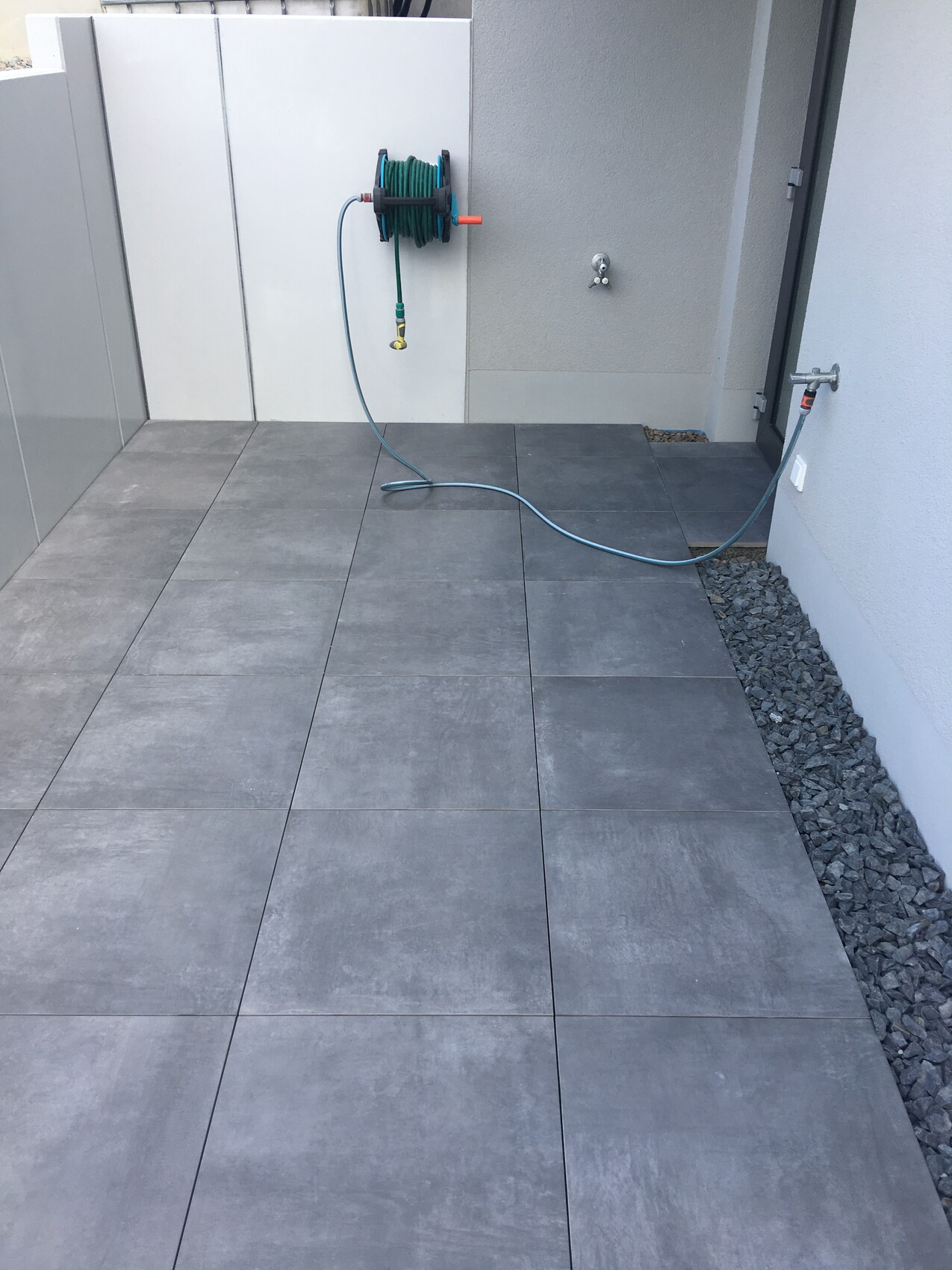 Graue Fliesen-Terrasse mit Gartenschlauch am Wandaufroller; Schlauch am Boden.