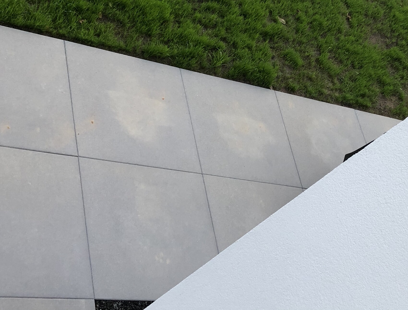 Außenbereich mit grauen Fliesen, Grasrand oben, diagonal verlaufende weiße Wand im Vordergrund