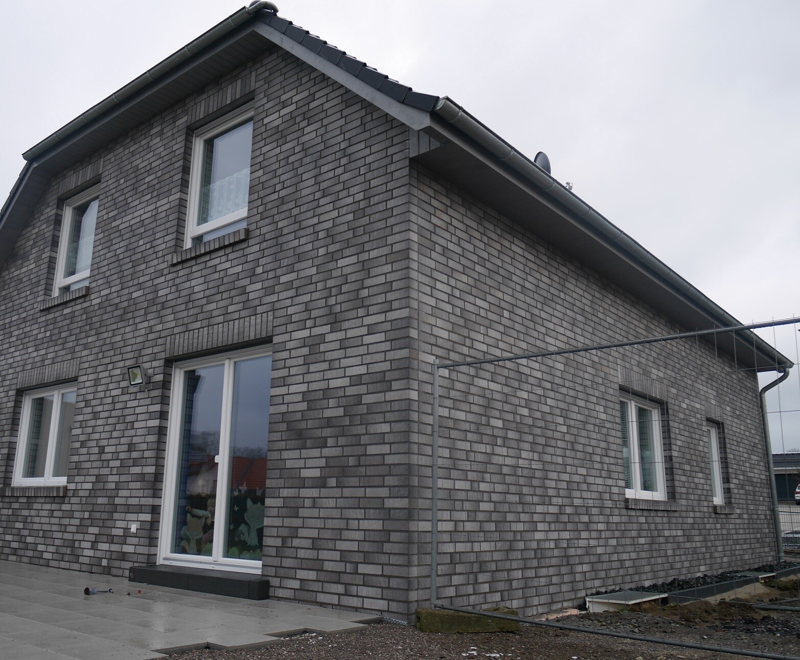 Graues Backsteinhaus mit Satteldach, mehreren Fenstern und Glas-Schiebetür im Erdgeschoss.