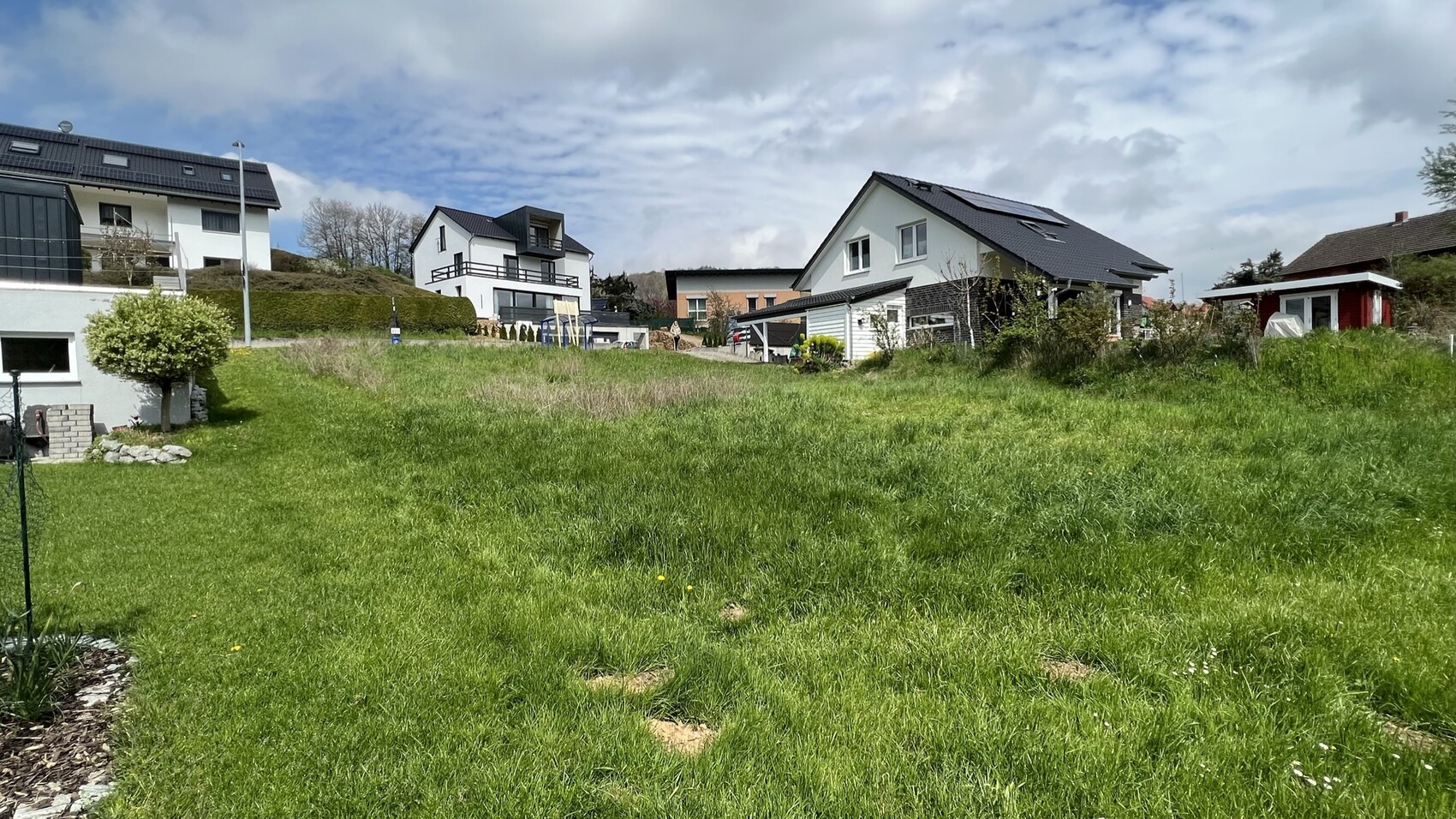 Großes grünes Grasfeld im Vordergrund, moderne Häuser am Hang, wolkiger Himmel.