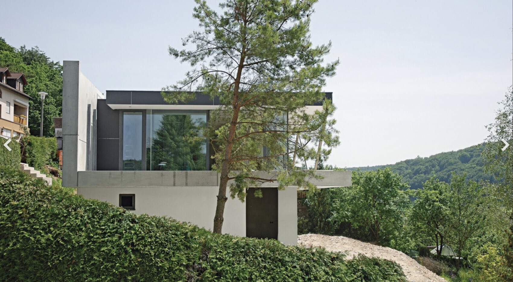 Moderne Beton-Glasvilla auf Hang mit großen Fenstern; Kiefer davor, umliegendes Grün.