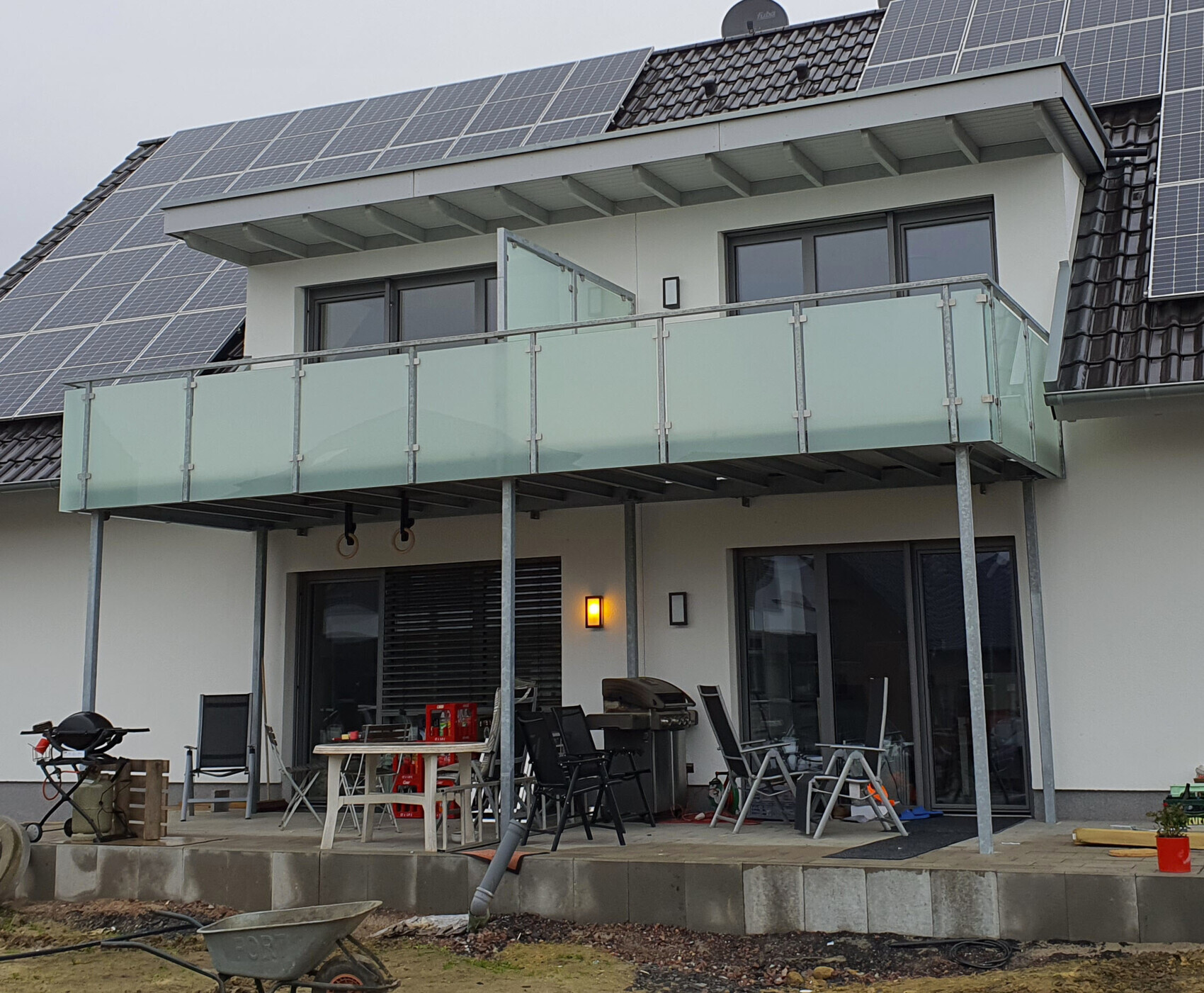 Moderne Hausfassade mit Balkon, Glasgeländer, Solaranlage und Terrasse mit Möbeln.