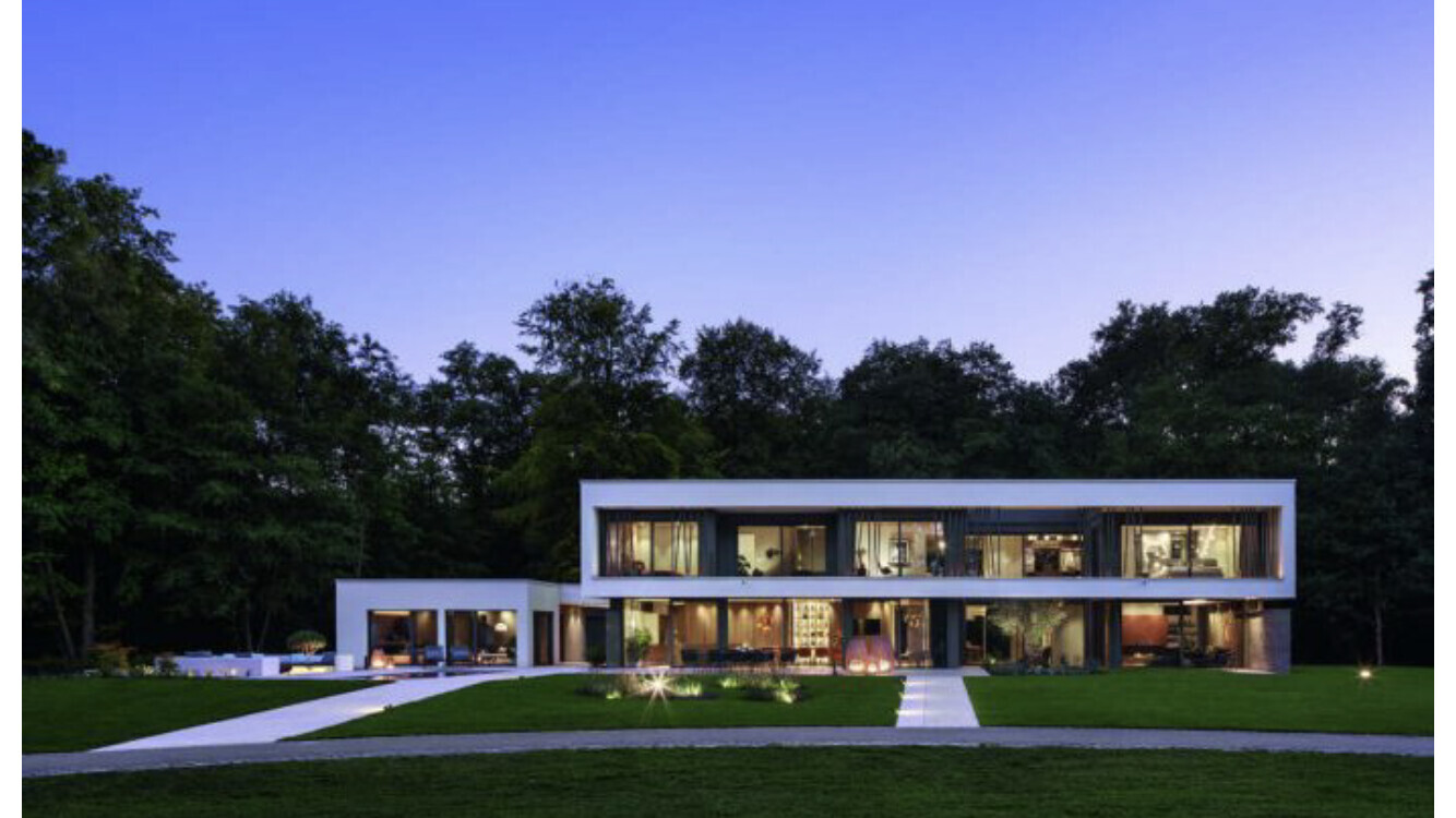 Moderne weiße Villa mit großer Glasfront, beleuchtetem Innenraum, grünem Garten.