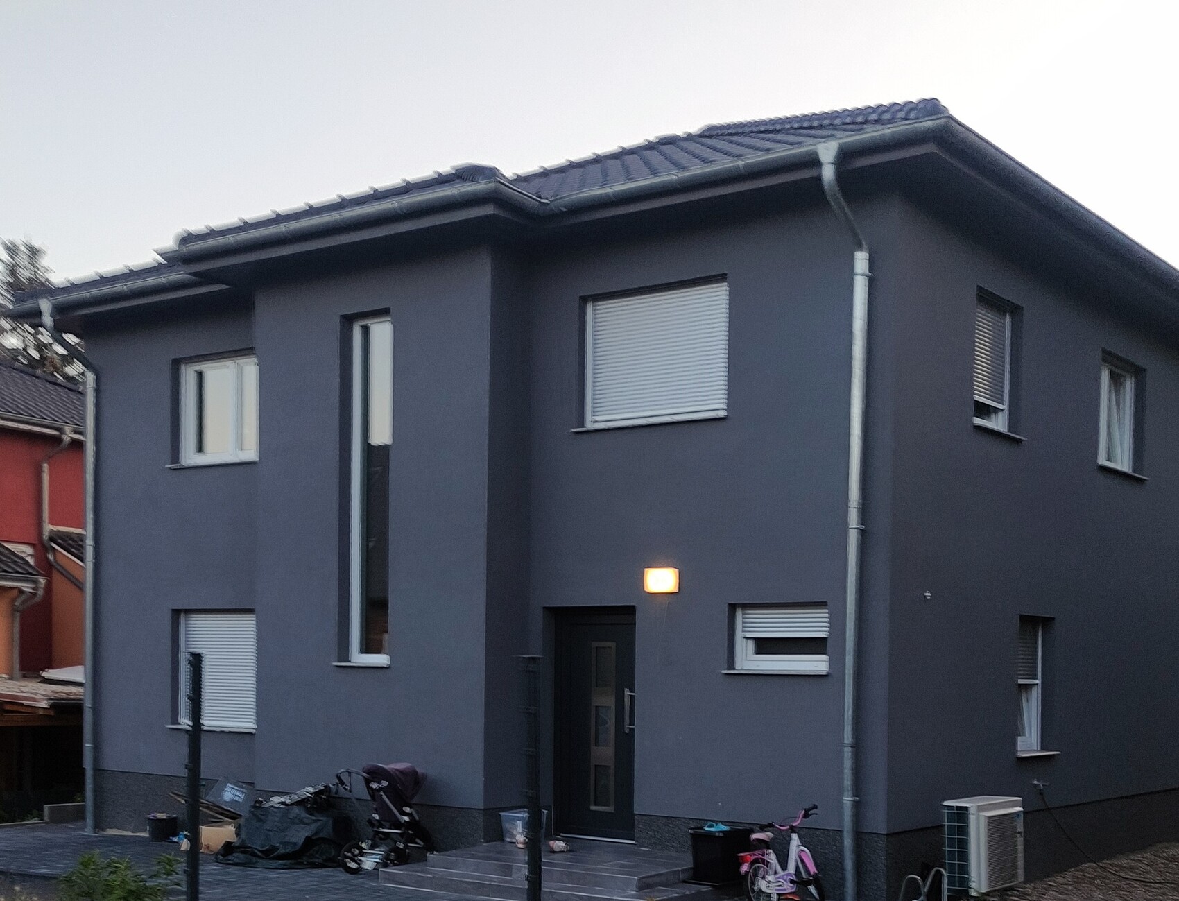 Modernes zweistöckiges graues Haus mit Fensterfront, Rollläden und beleuchteter Eingangstür.