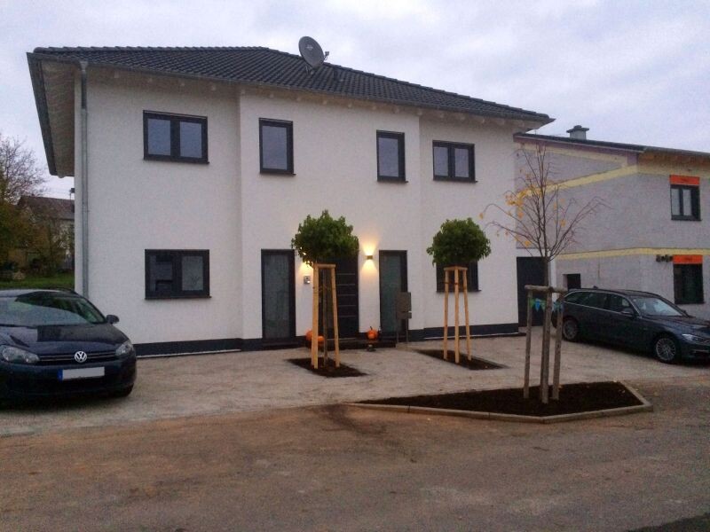 Zweistöckiges, weißes Wohnhaus mit Eingang, zwei gepflegte Bäume vorne, geparkte Autos.