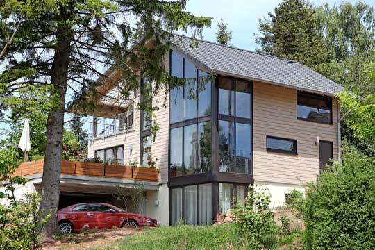 Modernes Holzhaus mit großen Glasfenstern, Balkon und rotem Auto vor Grünfläche.
