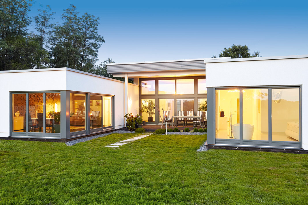 Modernes Haus mit Glasfronten, grünem Rasen, Steinpfad und Terrasse mit Sitzgruppe, warmes Innenlicht.