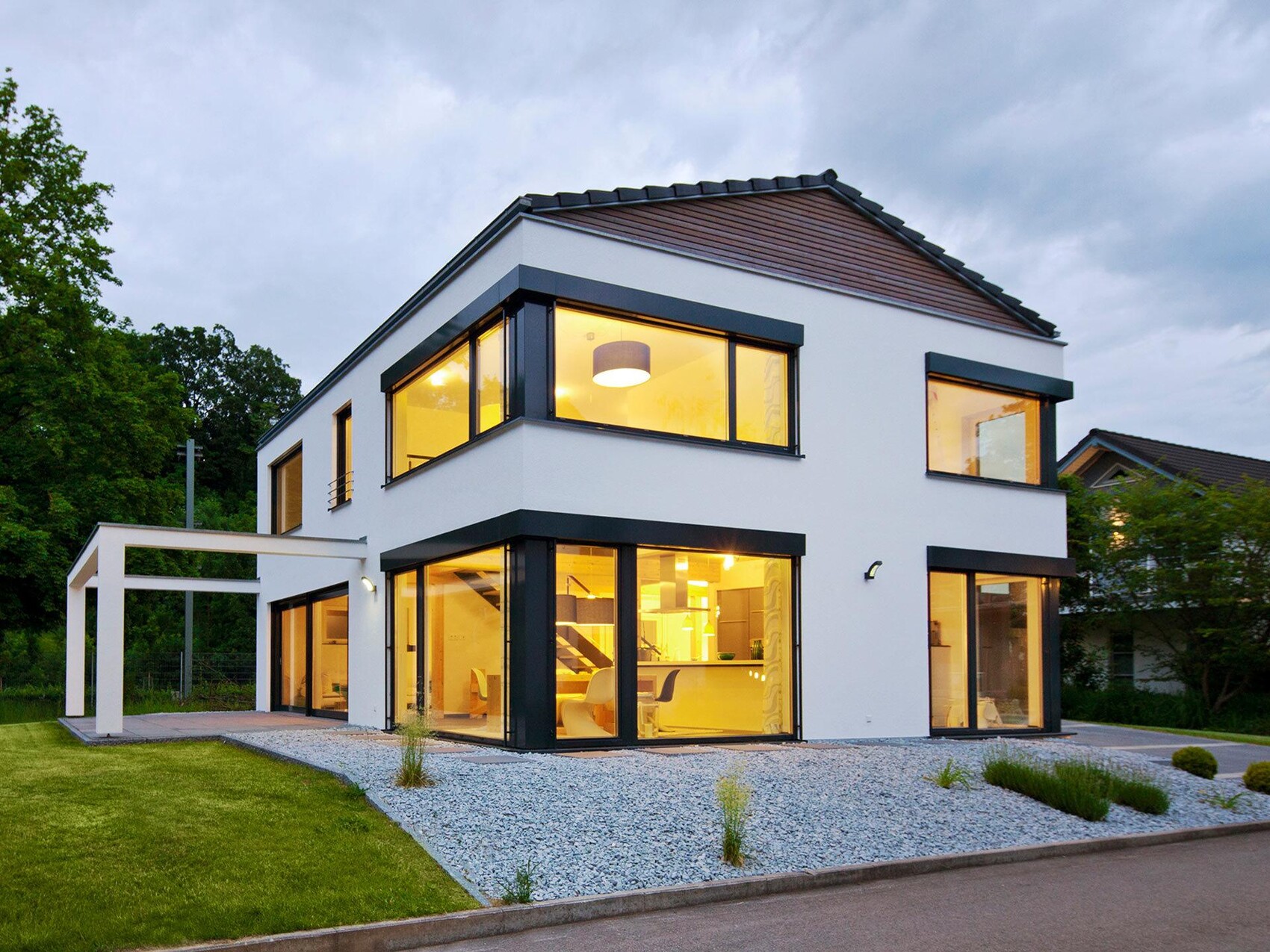 Moderne weiße Hausfassade mit großen Glasfronten, beleuchtete Innenräume bei Abendlicht.