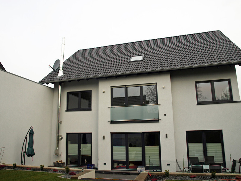 Modernes zweistöckiges Haus mit dunklem Ziegeldach, Glasbalkon, großen Fenstern und Garten.