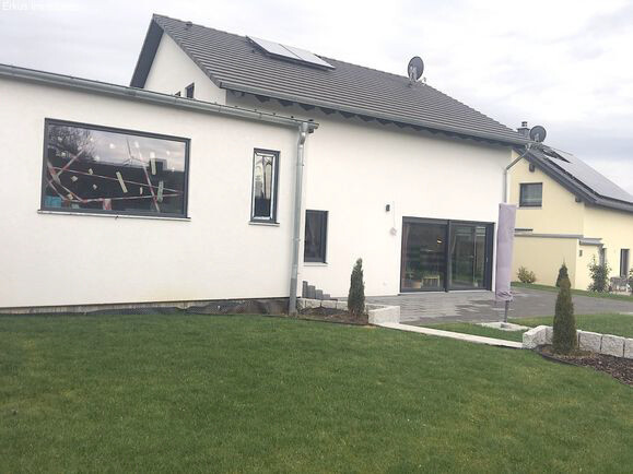Helles Einfamilienhaus mit Solarmodulen, großen Fenstern, gepflegtem Garten und Zufahrt.