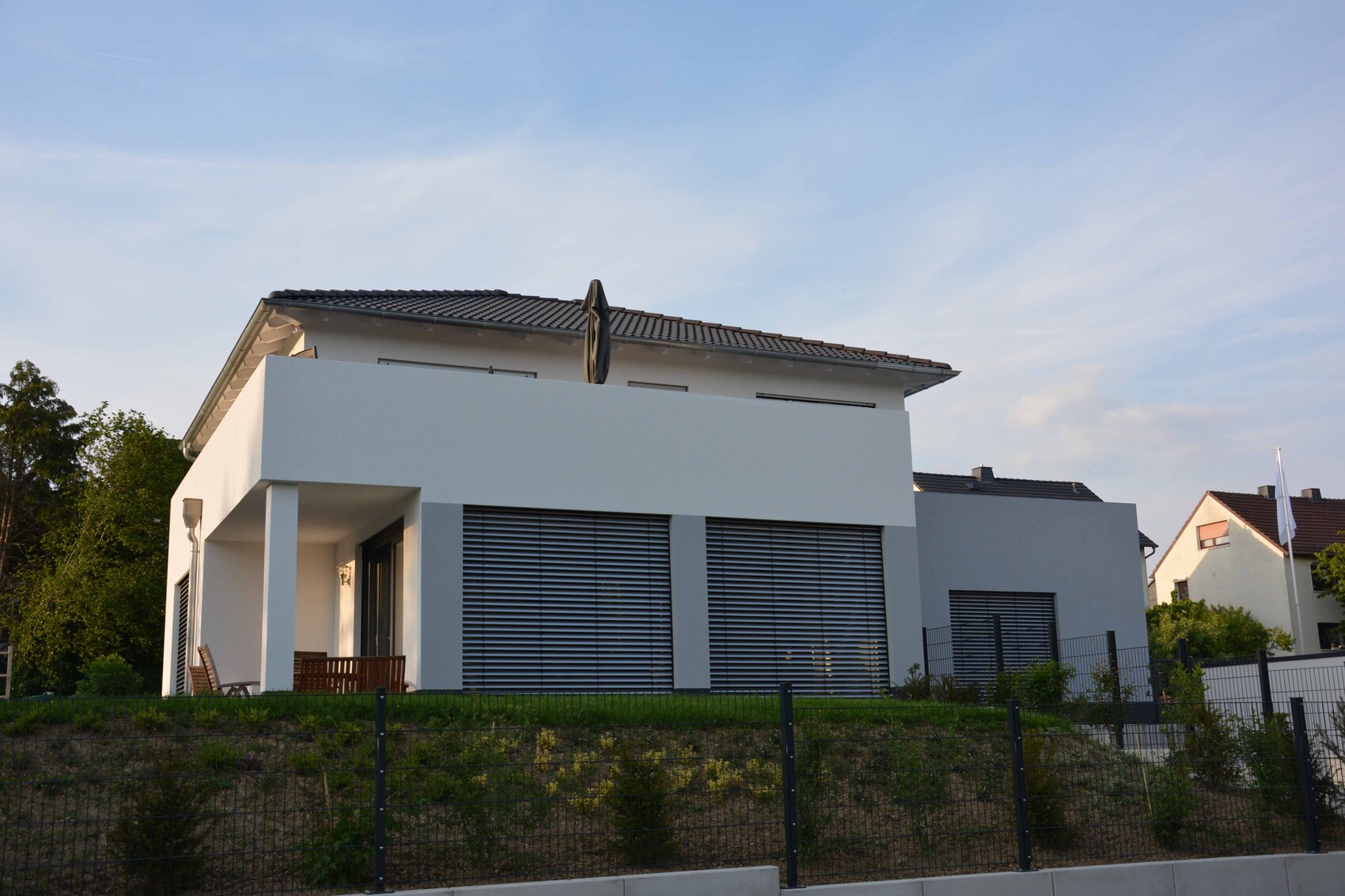 Modernes weißes Einfamilienhaus mit großen Fenstern, Rollläden, Terrasse und Gartenzaun.