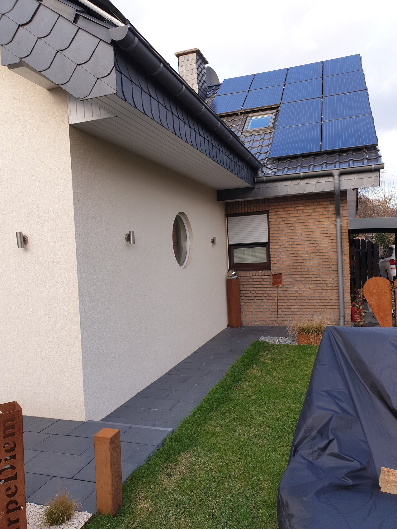Außenansicht eines Hauses mit blauen Solarmodulen auf dem Dach, rundem Fenster und Gartenweg.