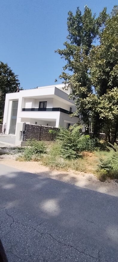 Moderner weißer zweistöckiger Wohnbau mit Balkon, Zaun und großen Bäumen am Rand.