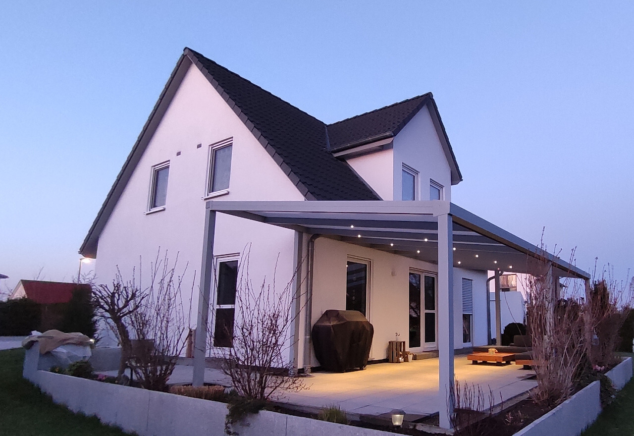 Ein modernes weißes Haus mit dunklem Dach, Pergola-Terrasse und Lichterketten.