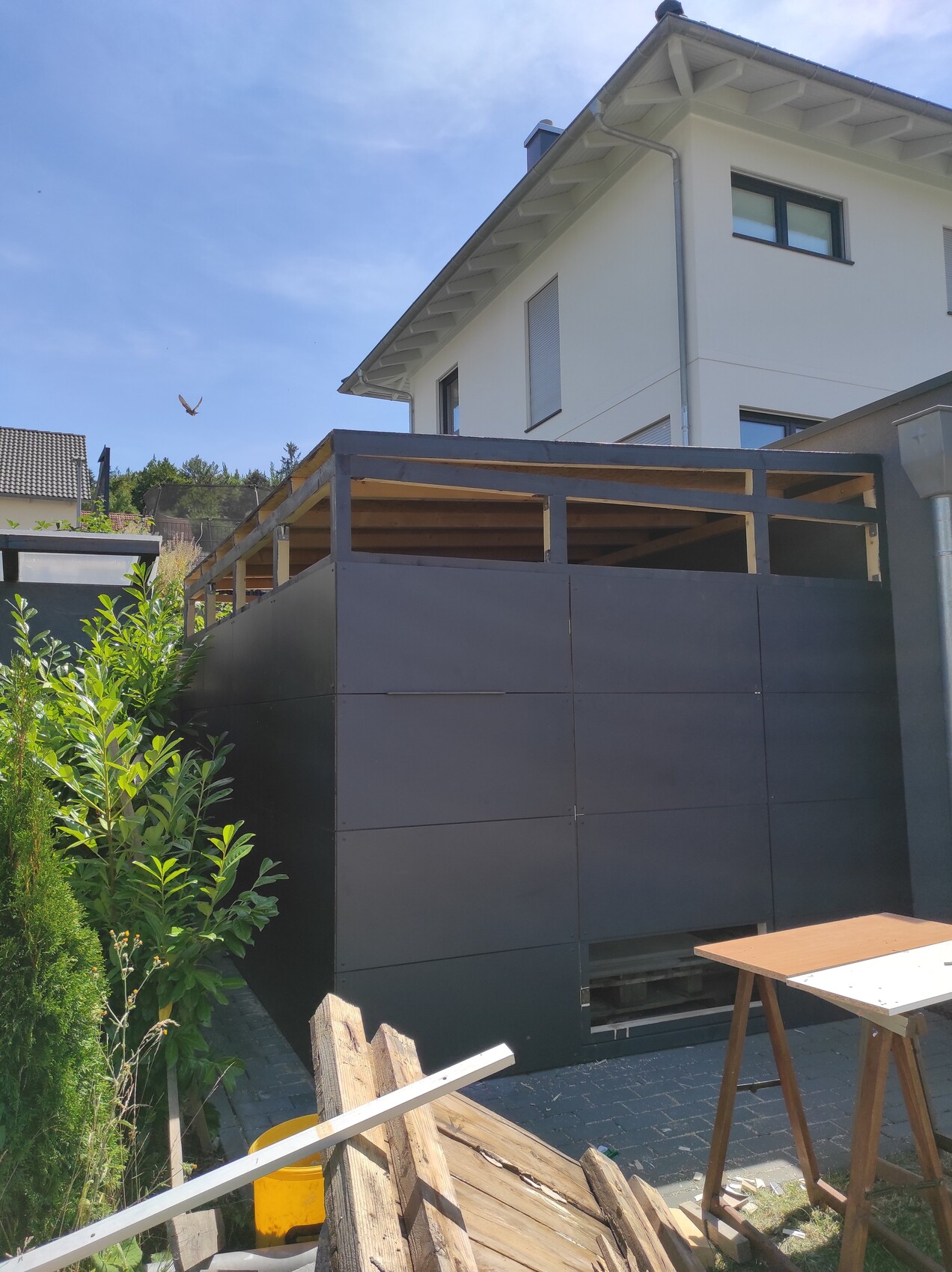 Weißes zweistöckiges Haus mit dunkler Metallwand und Holzdeck im Garten; Bauholz und Werkzeuge.