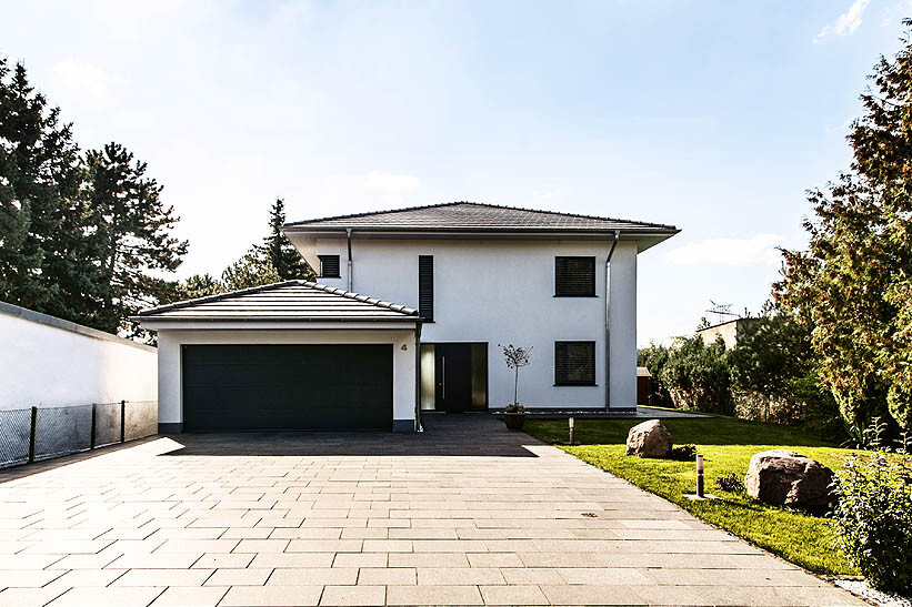 Modernes weißes Einfamilienhaus mit dunkler Garage, gepflasterter Zufahrt und Garten.