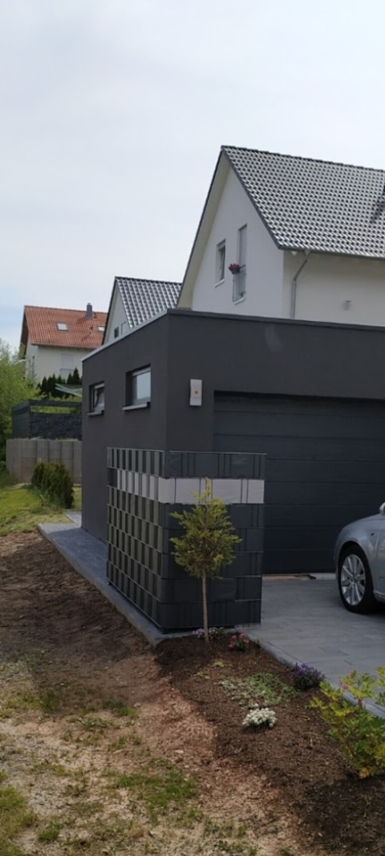 Moderne Doppelhaushälfte mit dunklem Garagenanbau, davor geparktes Auto und Garten mit Baum.
