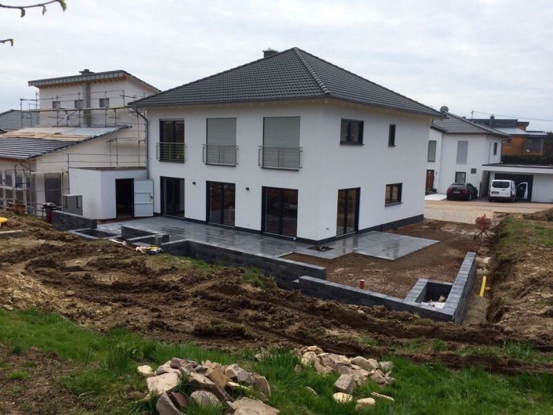 Modernes weißes Einfamilienhaus mit grauem Dach, Terrasse; Bauarbeiten und Erdhügel im Garten.