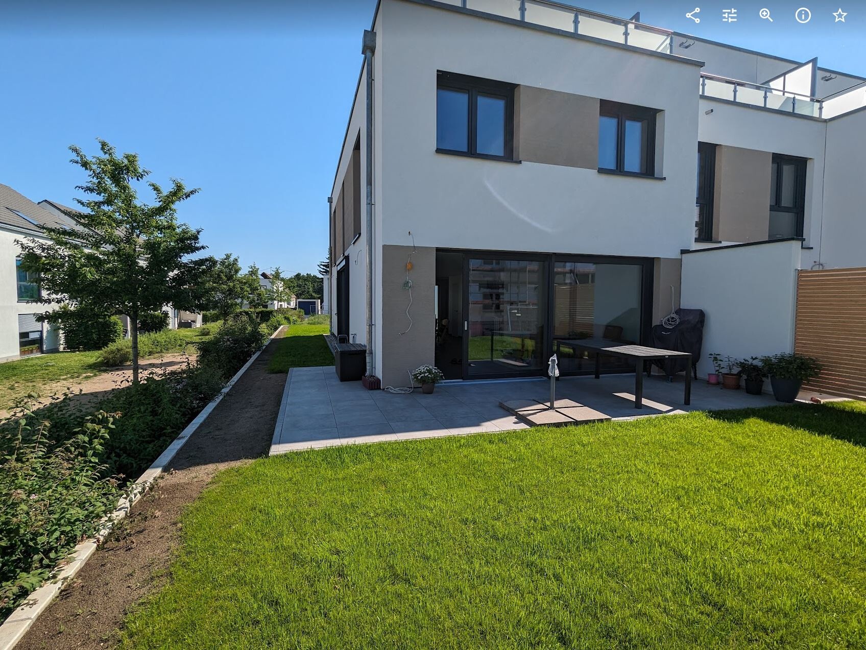 Modernes weißes Haus mit Terrasse, Glas-Schiebetür und grünem Rasen im Sonnenschein.