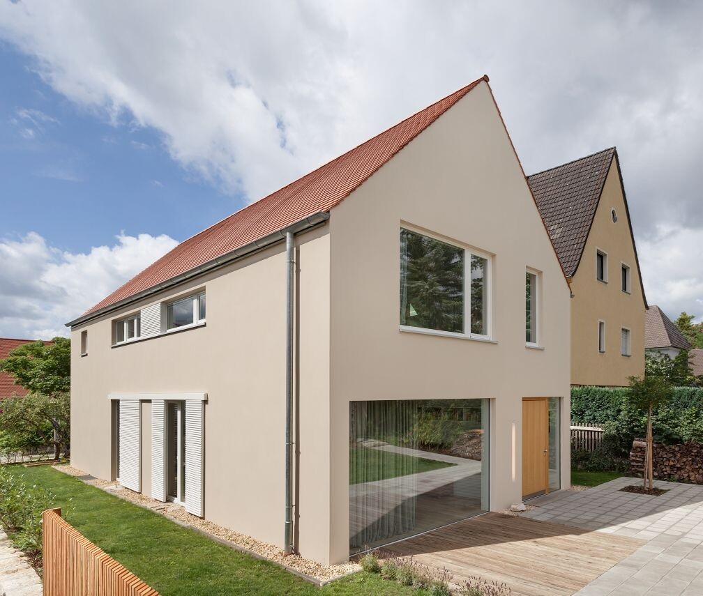 Modernes, helles Haus mit rotem Ziegeldach, großen Fenstern und Holzterrasse.