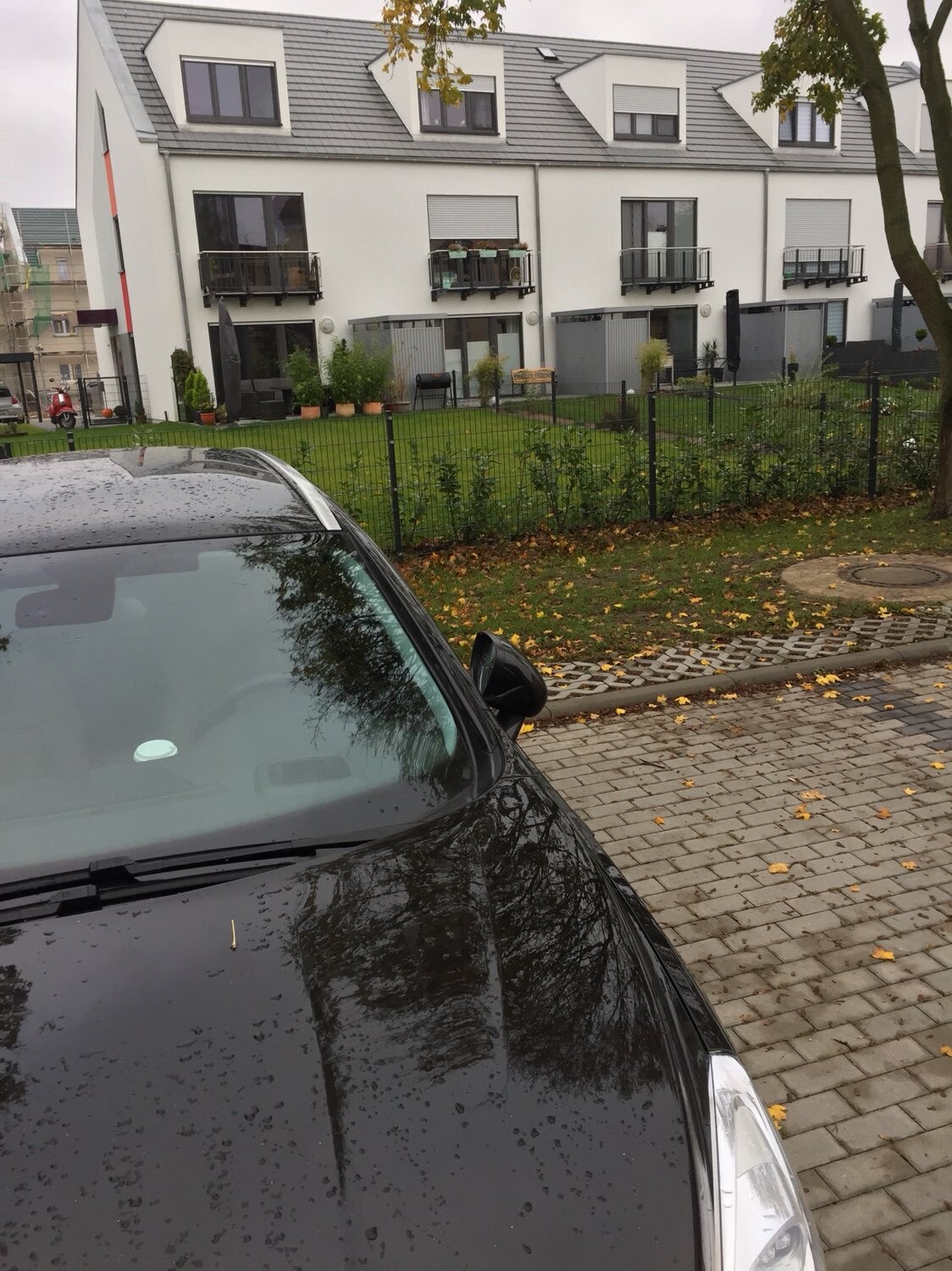 Schwarzer Wagen im nassen Vordergrund, weiße Mehrfamilienhäuser mit Balkonen hinter Gartenzaun.