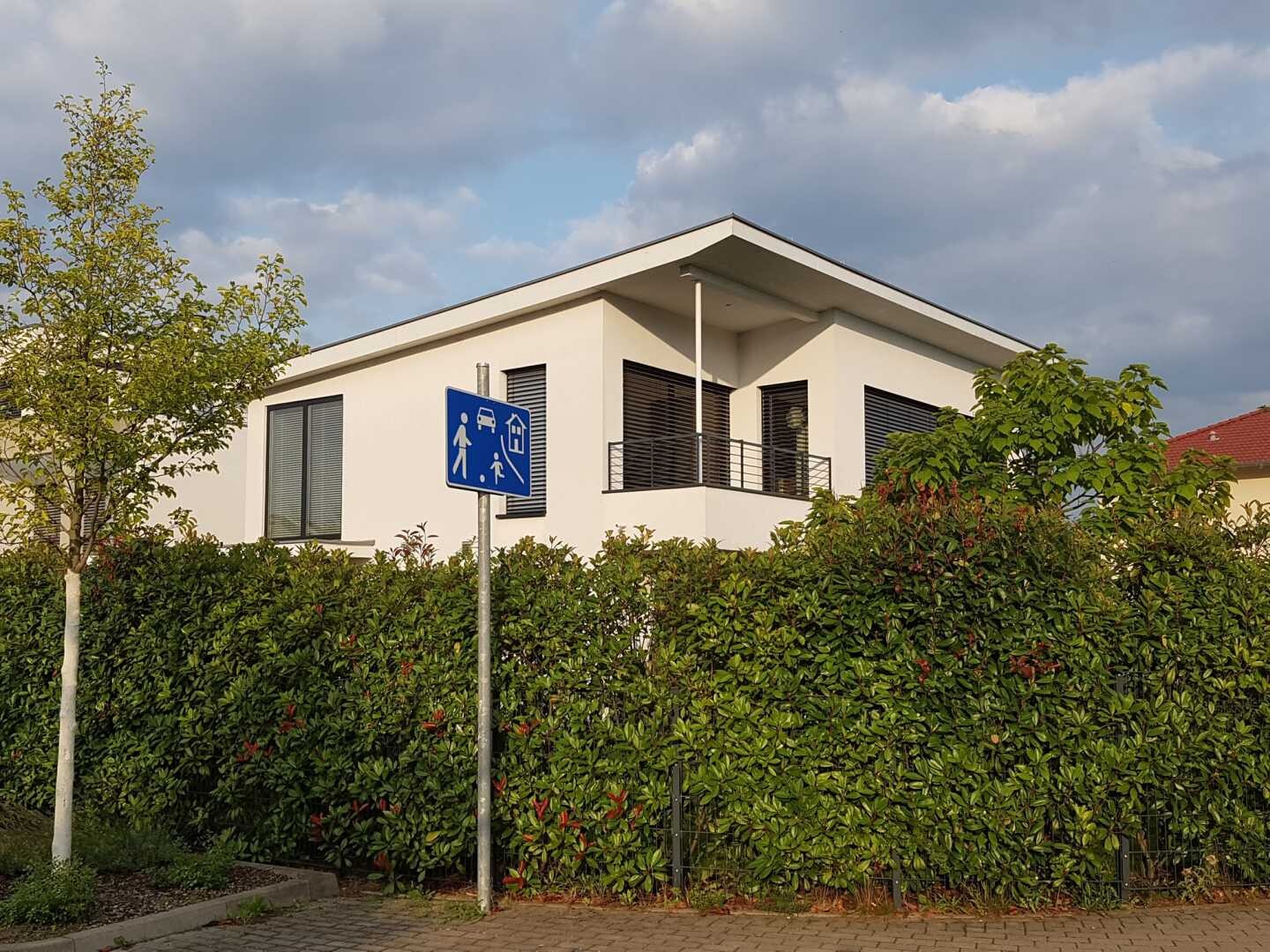 Modernes weißes Haus hinter Hecke; Balkon sichtbar, blaues Fußgänger-Schild am Straßenrand, bewölkt